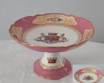 Könnte beinhalten: Eine Porzellanschale auf einem Fuß mit rosa Rand, goldenen Filigranverzierungen und einem weißen Becken, das mit einem farbigen Wappen verziert ist. Der Fuß ist weiß mit rosa Rand und einem Kronenmotiv, abgerundet durch Goldverzierungen.