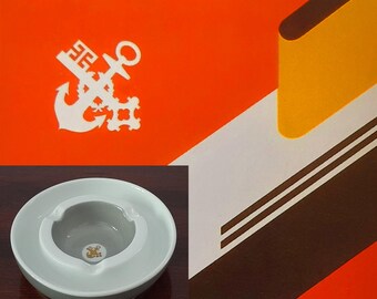 Könnte beinhalten: Ein weißer Keramik-Aschenbecher mit grauem Innenraum und goldenem Emblem. Der Aschenbecher befindet sich vor einem orange-weißen Grafikdesign mit dem Text "NORDDEUTSCHER LLOYD" und einem weißen Ankerlogo.