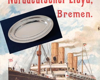 Könnte beinhalten: Eine silberne ovale Servierplatte mit doppeltem Rand, vor dunklem blauem Hintergrund. Die Platte wird über einer Vintage-Illustration eines Schiffes mit dem Text "Norddeutscher Lloyd, Bremen" präsentiert.