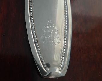 Könnte beinhalten: Ein silberfarbener Löffelstiel mit einer glatten, reflektierenden Oberfläche, die von einem Perlenrand gesäumt ist. In der Mitte des Stiels befindet sich ein eingraviertes Wappen mit den Buchstaben B.A.C. und einem kleinen Ankersymbol.