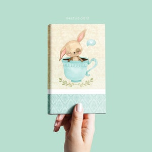May include: A rectangular canvas print featuring a whimsical illustration of a bunny nestled inside a light blue teacup. The bunny has closed eyes and long ears. The teacup is decorated with a floral design. The background is a light beige with a floral pattern.