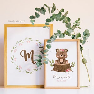 May include: Two framed prints with a woodland theme. The first print features a large letter "M" surrounded by a wreath of green leaves and pink flowers. The second print features a brown bear wearing a pink flower crown sitting on a tree stump with pink flowers. The text "Manuela 03/11/2018" is printed below the bear.