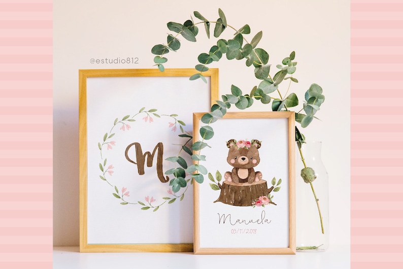 May include: Two framed prints with a woodland theme. The first print features a large letter "M" surrounded by a wreath of green leaves and pink flowers. The second print features a brown bear wearing a pink flower crown sitting on a tree stump with pink flowers. The text "Manuela 03/11/2018" is printed below the bear.