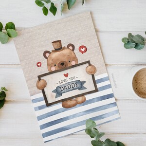 Puede incluir: Una ilustraci&oacute;n de acuarela de un oso marr&oacute;n con un sombrero y sosteniendo un cartel que dice "Te quiero pap&aacute;". El oso est&aacute; sobre un fondo de rayas azules y blancas.