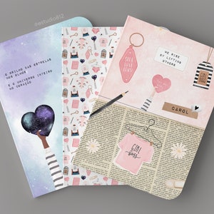 May include: Three pink and white notebooks with different designs. The first notebook has a galaxy design with the text "O brilho das estrelas nos olhos e o universo inteiro no cora&ccedil;&atilde;o". The second notebook has a pink and white pattern with hearts, keys, and other small objects. The third notebook has a pink background with a newspaper print and the text "We rise by lifting others" and "Girl Boss".
