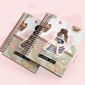 Op de afbeelding: Twee bruine spiraalgebonden planners met een vintage wereldkaartontwerp. De voorkant heeft een cartoonillustratie van een vrouw met bruin haar die een foto maakt met een camera. De tekst op de omslag luidt "Wanderlust Planner 2021".