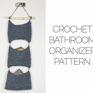 以下が含まれることがあります： くすんだ青灰色の3ポケットのかぎ針編みバスルームオーガナイザー。木のダボから吊り下げられています。オーガナイザーは金属製のブラケットで壁に取り付けられています。「CROCHET BATHROOM ORGANIZER PATTERN」の文字が見えます。