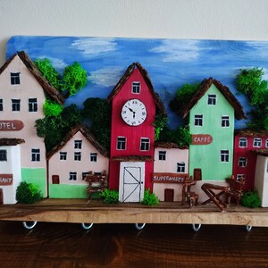 May include: A wooden key holder shaped like a small town with five buildings, including a hotel, restaurant, cafe, supermarket, and a clock tower. The buildings are painted in various colours, including pink, green, red, and white. The key holder has a rustic wooden base and metal hooks for hanging keys.