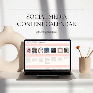 Puede incluir: Un portátil que muestra un calendario de contenido de redes sociales rosa y blanco. El texto "SOCIAL MEDIA CONTENT CALENDAR" y "@thebloggingblond" son visibles. Un jarrón beige y un portalápices con pinceles están sobre la superficie blanca.