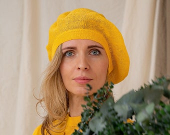 May include: A woman wearing a yellow beret and a yellow sweater holds a bouquet of green eucalyptus branches.