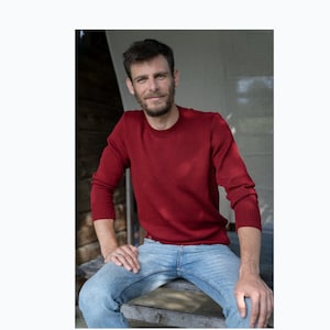 Peut inclure: Un homme portant un pull rouge et un jean bleu est assis sur un banc en bois. Il sourit et regarde la caméra.