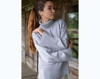 May include: A light gray long-sleeve sweater featuring a high turtleneck, wide ribbed cuffs, and a ribbed hem with side slits. The knit garment has a textured finish and a small rectangular label attached to the bottom hem near the side opening.