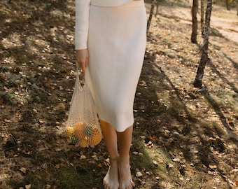 May include: A woman wearing a white knitted sweater and skirt set stands in a forest. She is holding a mesh bag filled with lemons.