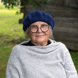 Puede incluir: Una mujer con un poncho de punto gris y una boina azul con gafas. Está sentada en una silla con las manos juntas en su regazo.