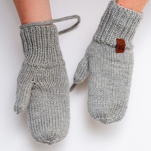 Pode incluir: Um par de luvas de malha cinzas com uma etiqueta de couro marrom que diz "NIGE". As luvas têm um punho dobrável e um design sem dedos.