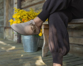 May include: A pair of light brown suede clogs with a buckle closure. The clogs are worn by a person sitting on a wooden bench.