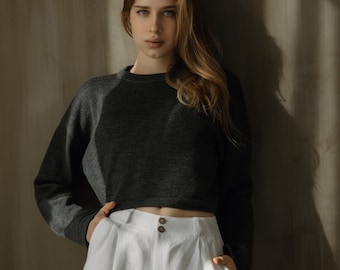May include: A woman wearing a dark gray cropped sweater and white linen shorts. She is standing against a light brown wall with shadows from the sun.