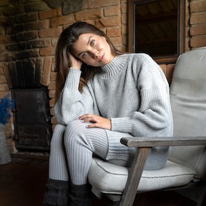K&ouml;nnte beinhalten: Eine Frau in einem grauen Strickpullover und einer passenden Hose sitzt auf einem Holzstuhl mit einem wei&szlig;en Kissen. Sie tr&auml;gt schwarze Stricksocken und hat ihre Hand auf ihrem Bein. Der Stuhl steht vor einer Backsteinwand mit einem Spiegel an der Wand.