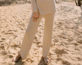 May include: A woman wearing a light beige knitted sweater and matching trousers. The outfit is ribbed and has a relaxed fit. She is standing in a sandy area with trees in the background.