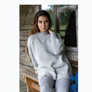 K&ouml;nnte beinhalten: Ein grauer Strickpullover mit hohem Kragen und langen &Auml;rmeln. Der Pullover ist locker geschnitten und hat eine gerippte Struktur. Das Model tr&auml;gt den Pullover mit grauen Leggings.