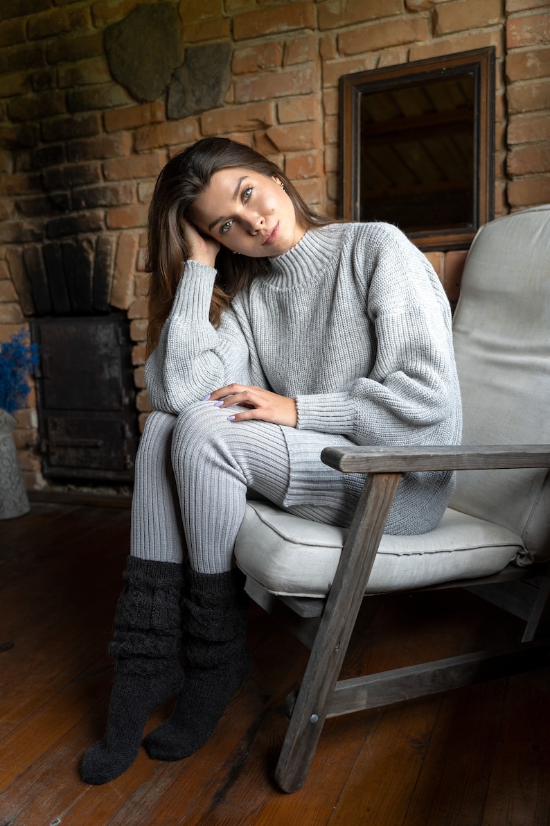 K&ouml;nnte beinhalten: Eine Frau in einem grauen Strickpullover und einer passenden Hose sitzt auf einem Holzstuhl mit einem wei&szlig;en Kissen. Sie tr&auml;gt schwarze Stricksocken und hat ihre Hand auf ihrem Bein. Der Stuhl steht vor einer Backsteinwand mit einem Spiegel an der Wand.