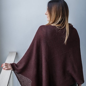 May include: A woman wearing a maroon knitted poncho with a long, straight hairstyle. She is standing in front of a white wall and holding onto a white railing.