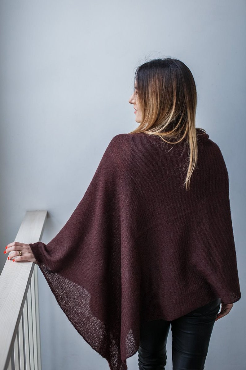 May include: A woman wearing a maroon knitted poncho with a long, straight hairstyle. She is standing in front of a white wall and holding onto a white railing.