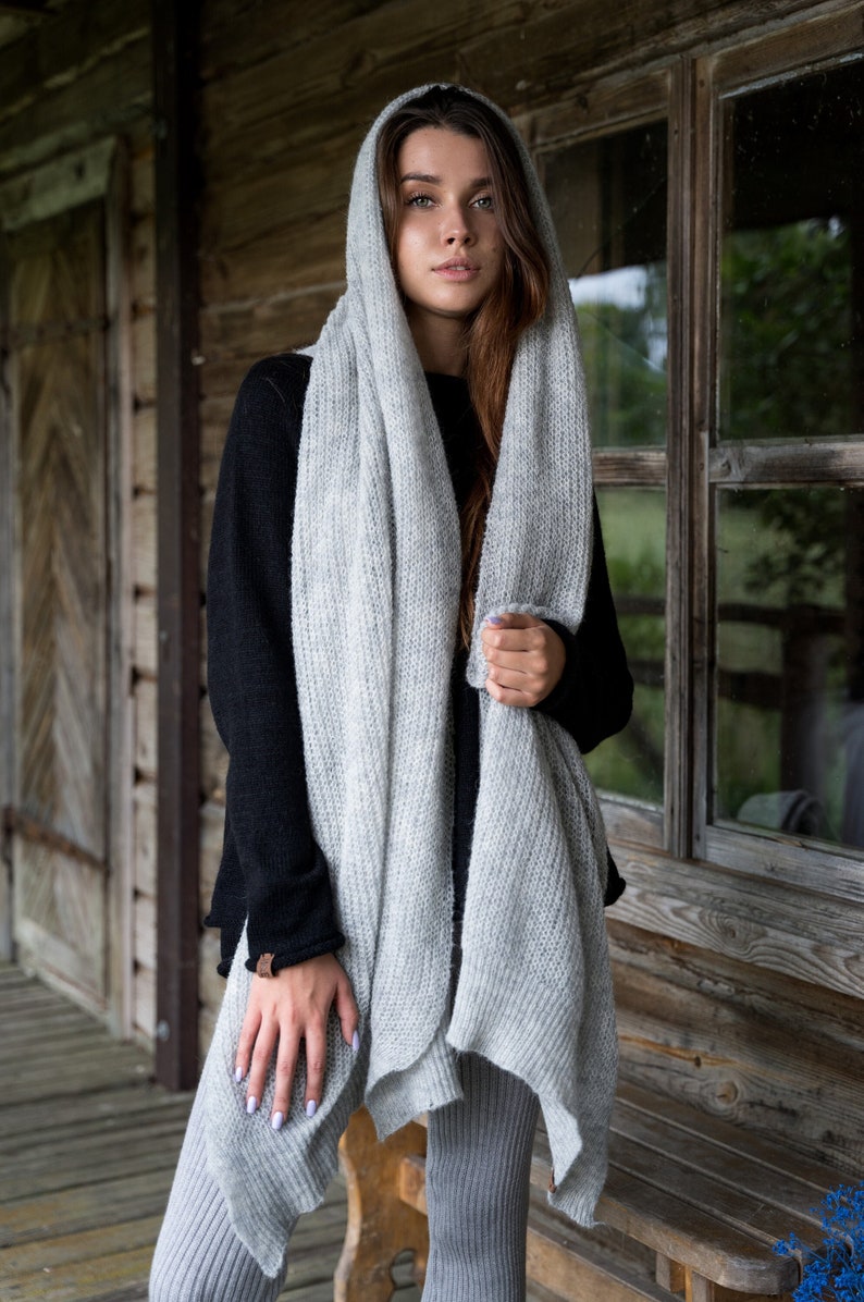 May include: A woman wearing a gray knitted scarf draped over her head and shoulders. The scarf is a light gray color and has a loose, flowing texture. The woman is standing in front of a wooden building with a window behind her.