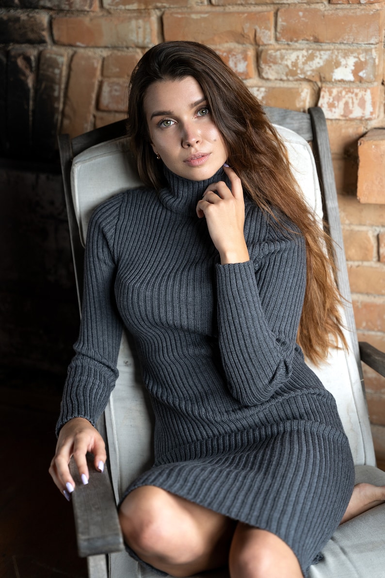May include: A woman wearing a gray ribbed turtleneck sweater dress, sitting in a chair with a white cushion. She is looking at the camera with her hand on her chin.