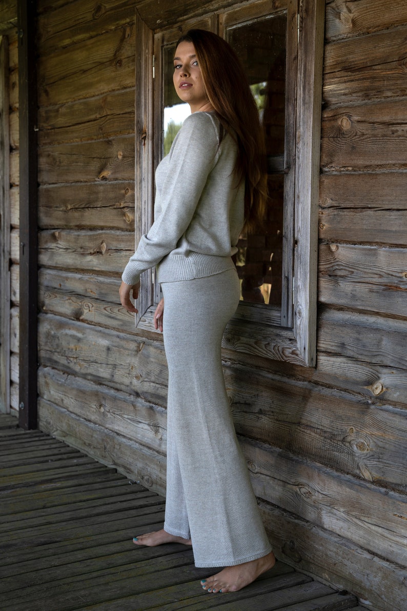 May include: A woman wearing a light gray knit sweater and matching pants stands in front of a wooden wall. She is looking over her shoulder at the camera.