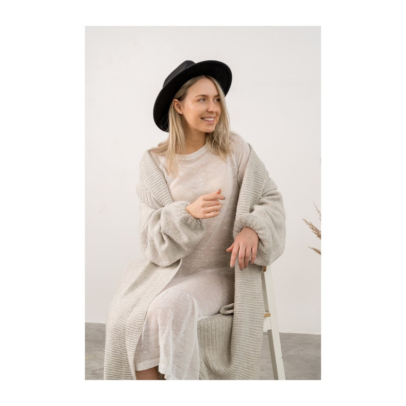 May include: A woman wearing a light gray cardigan over a white sheer dress. She is wearing a black fedora and sitting on a white chair.