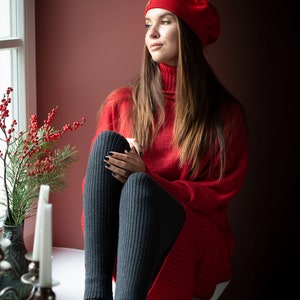 May include: A woman wearing a red sweater and a red beret sits by a window. She is wearing black tights and gray slippers with colorful embroidery.