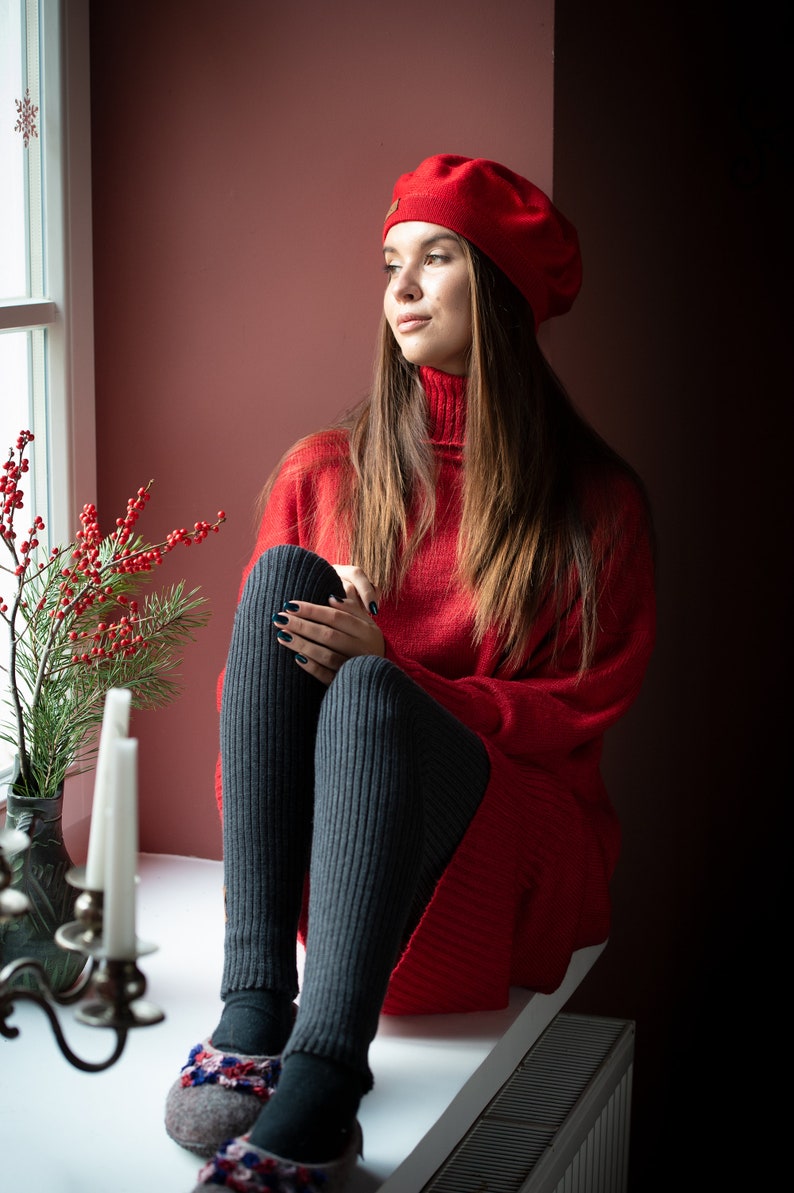 May include: A woman wearing a red sweater and a red beret sits by a window. She is wearing black tights and gray slippers with colorful embroidery.