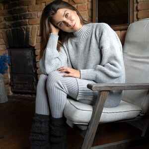 May include: A woman wearing a gray knitted sweater and matching pants sits in a wooden chair with a white cushion. She is wearing black knitted socks and has her hair pulled back from her face.