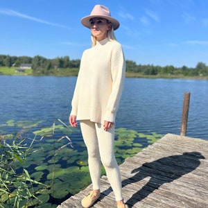 May include: A woman wearing a white knitted sweater and matching pants with a wide leg. She is wearing a light pink fedora hat and brown leather shoes. She is standing on a wooden dock over a lake with lily pads.