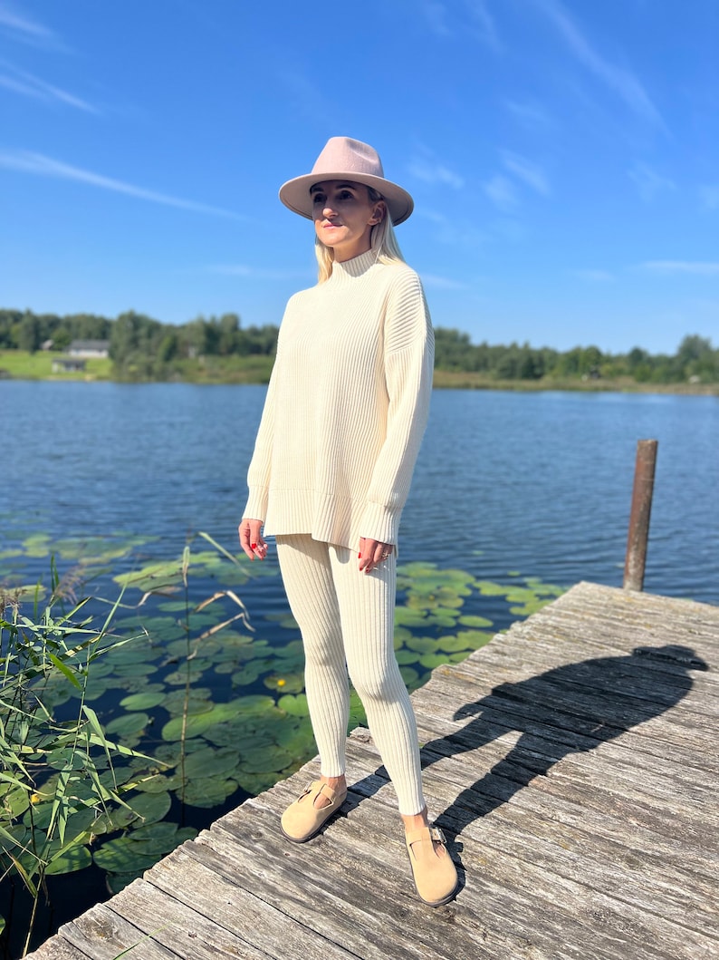 May include: A woman wearing a white knitted sweater and matching pants with a wide leg. She is wearing a light pink fedora hat and brown leather shoes. She is standing on a wooden dock over a lake with lily pads.