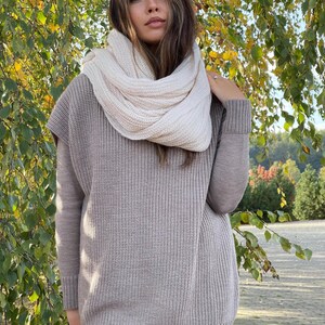 May include: A beige knitted sweater vest with long sleeves and a white knitted scarf. The sweater has a loose fit and a ribbed texture.