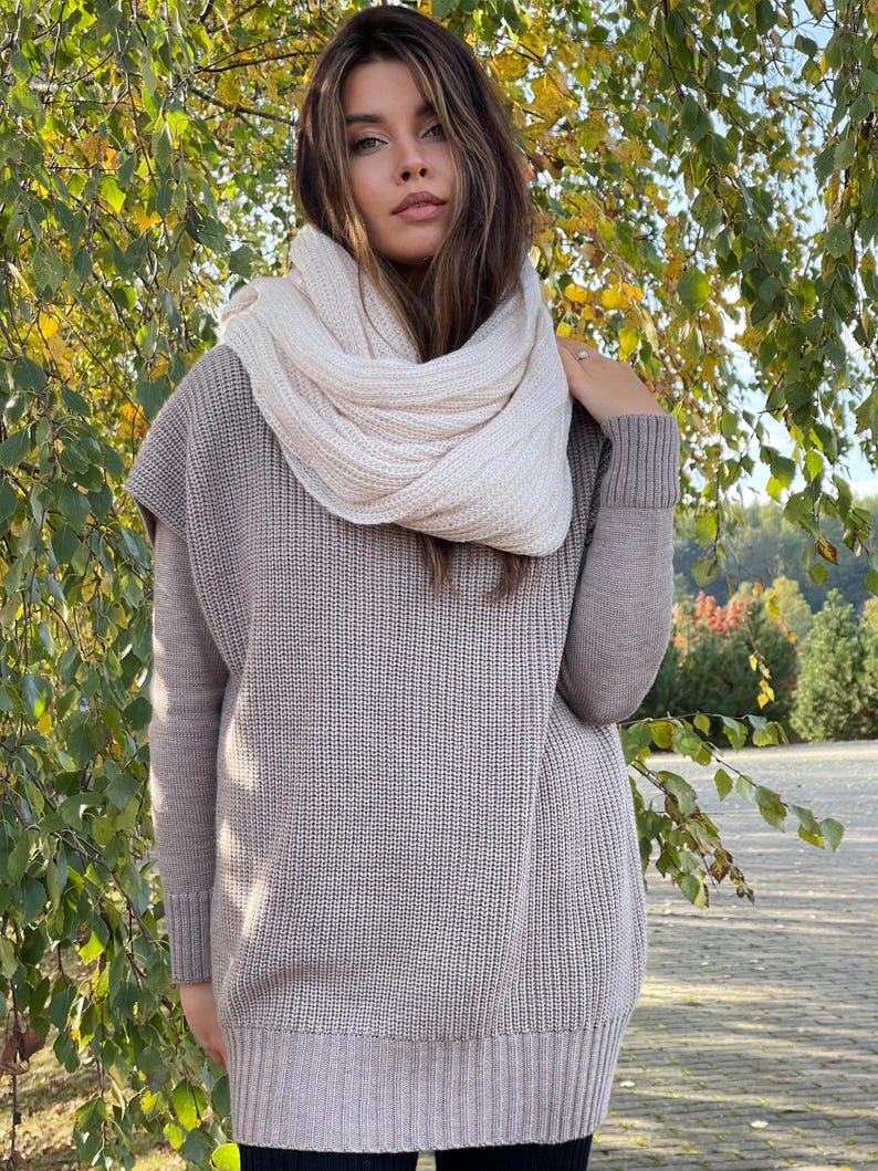 May include: A beige knitted sweater vest with long sleeves and a white knitted scarf. The sweater has a loose fit and a ribbed texture.