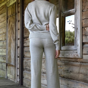 May include: A woman wearing a light gray sweater and matching pants stands in front of a wooden wall with a window. The sweater has a v-neck and long sleeves. The pants are wide-legged and have a relaxed fit.