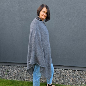 May include: A woman wearing a grey poncho with a blue denim shirt and black sandals. She is standing in front of a grey wall and green grass.
