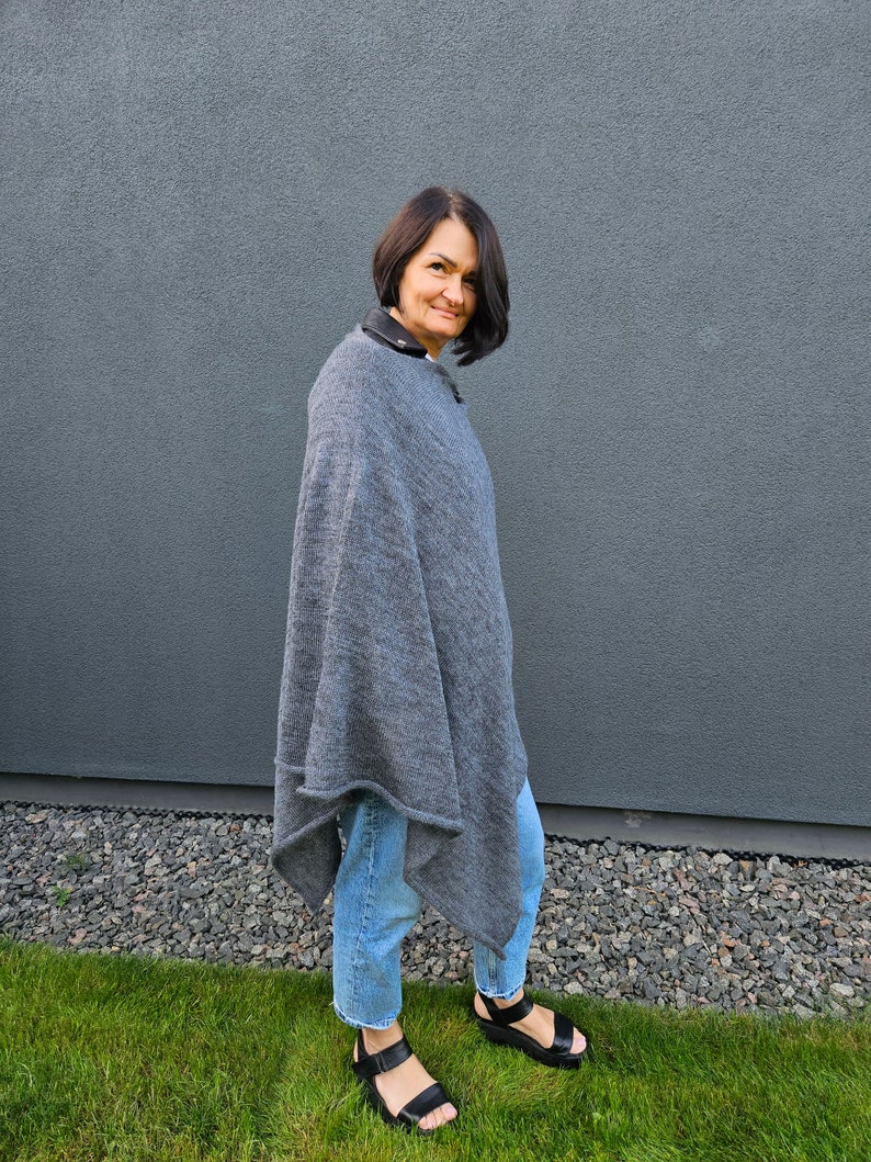 May include: A woman wearing a grey poncho with a blue denim shirt and black sandals. She is standing in front of a grey wall and green grass.
