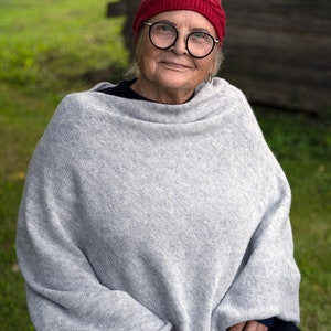 May include: A woman wearing a grey knitted poncho with a red knitted headband and glasses. She is sitting outdoors in front of a wooden wall.
