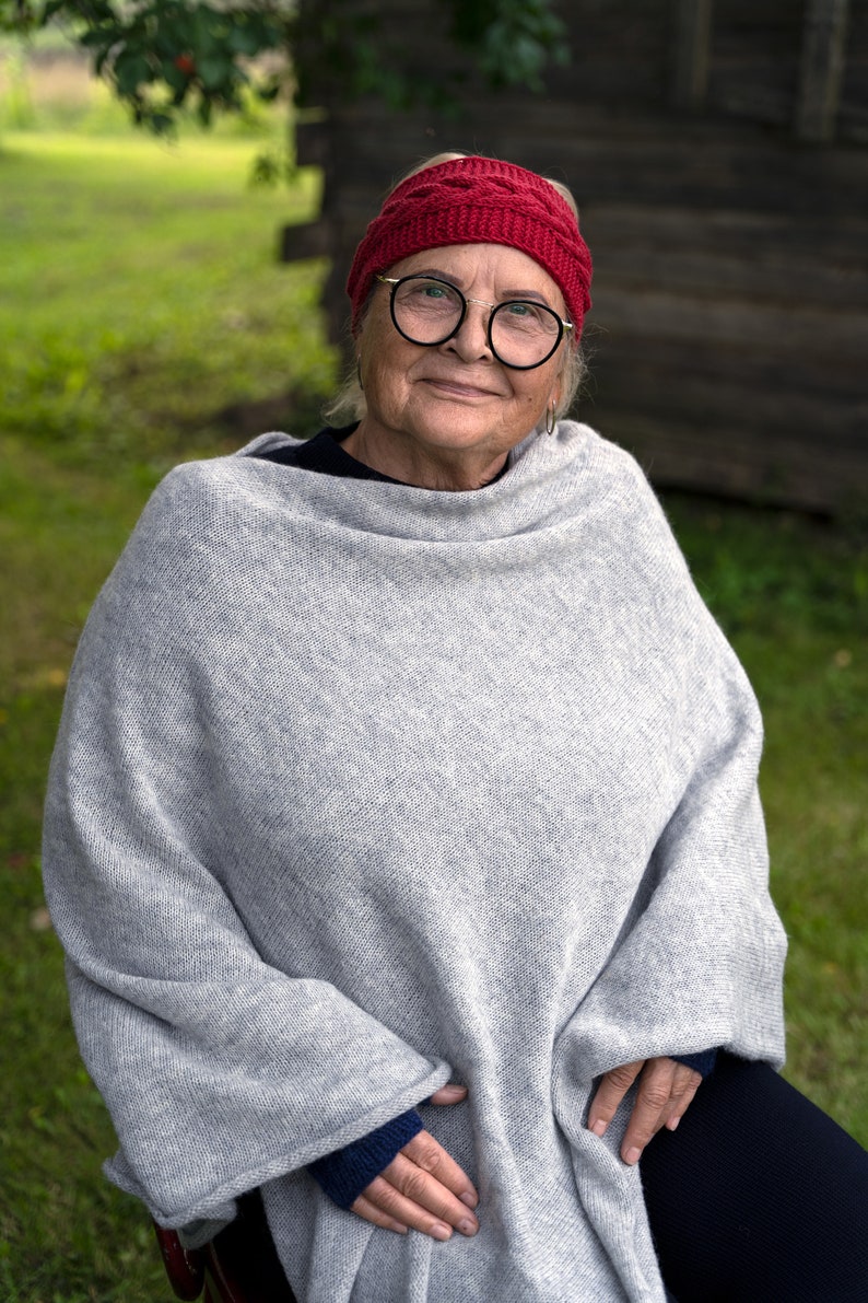 May include: A woman wearing a grey knitted poncho with a red knitted headband and glasses. She is sitting outdoors in front of a wooden wall.