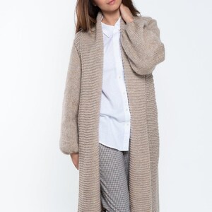 May include: A long, beige cardigan with a shawl collar and long sleeves. The cardigan is knitted with a textured pattern and has a relaxed fit.