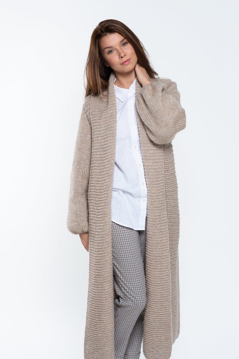 May include: A long, beige cardigan with a shawl collar and long sleeves. The cardigan is knitted with a textured pattern and has a relaxed fit.