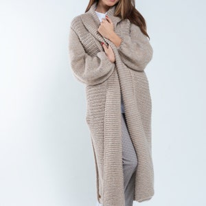 May include: A long, beige knitted cardigan with a shawl collar. The cardigan is worn over a white t-shirt and plaid pants. The model is wearing white sneakers.