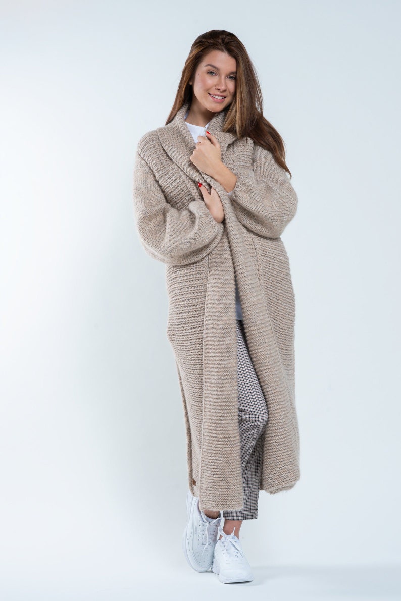 May include: A long, beige knitted cardigan with a shawl collar. The cardigan is worn over a white t-shirt and plaid pants. The model is wearing white sneakers.