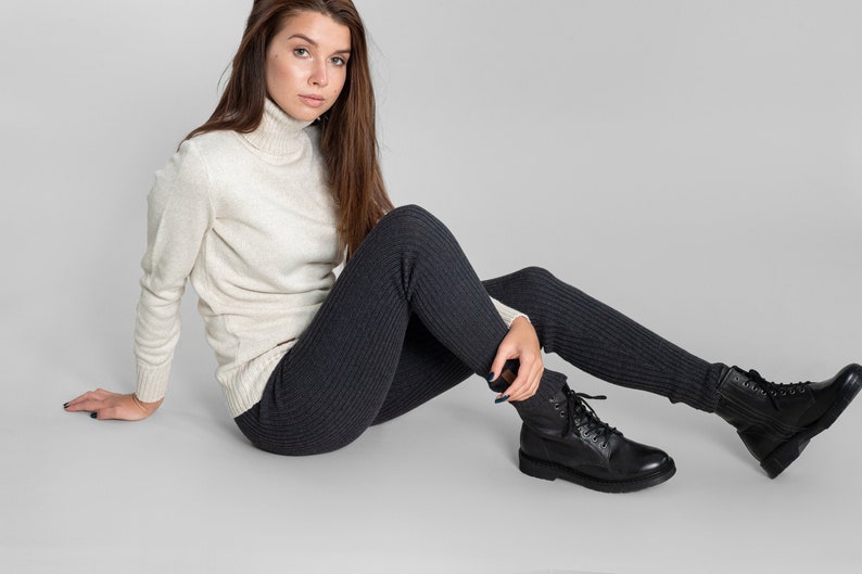 May include: A woman wearing a white turtleneck sweater and dark gray ribbed leggings. She is sitting on a gray background and wearing black lace-up boots.