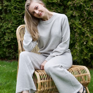 May include: A woman wearing a light gray knit sweater and matching pants sits on a wicker chair. She is wearing black sandals and has long blonde hair.