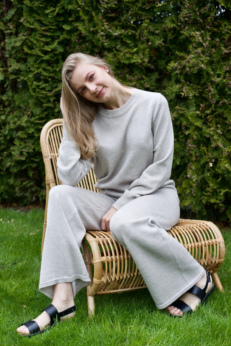 May include: A woman wearing a light gray knit sweater and matching pants sits on a wicker chair. She is wearing black sandals and has long blonde hair.